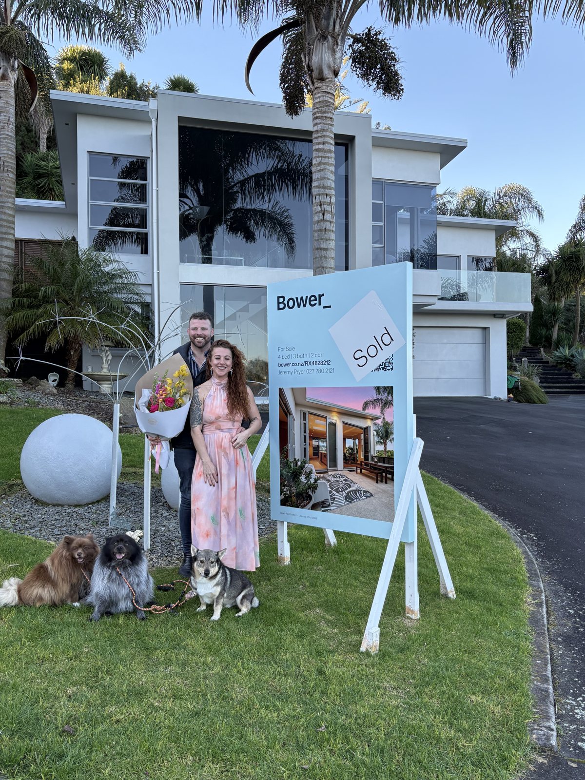 Dani and Alex beside a Sold sign outside their new home with their dogs